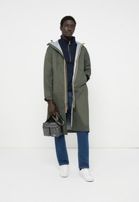 Giacca lunga impermeabile verde con foderatura grigia, accenti arancioni e gialli. Indossata sopra una maglia blu navy con zip, insieme a jeans blu e sneakers bianche. Tiene una borsa grigia.