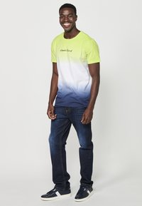 T-shirt s kratkim rukavima u gradijentu od limeta zelene do tamnoplave, s okruglim izrezom i crnim izvezenim logom, u kombinaciji s tamnoplavim trapericama i tenisicama.