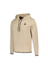 Sudadera beige con un bolsillo delantero, cordones negros y un logo en el pecho. Textura suave con un corte relajado y acentos negros contrastantes.