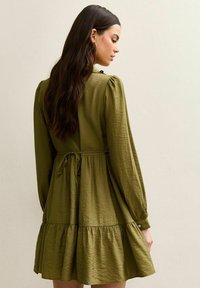 Olijfgroene jurk met een gelaagde gerimpelde zoom, lange mouwen en een detail met strik in de taille. Gestructureerde stof met een licht gekreukte uitstraling.