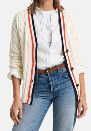 Person iført en cremefarvet kabelstrikket cardigan med røde, hvide og sorte kanter, ærmerne smøget op, hvid skjorte med knapper, brunt bælte og blå jeans.