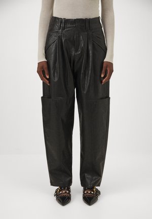 Personne portant un pantalon en cuir noir taille haute avec de grandes poches et des chaussures noires pointues avec des brides cloutées.