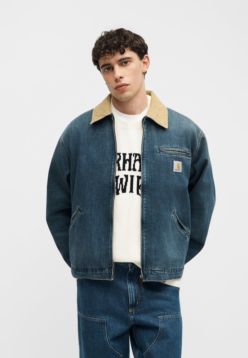 Giovane uomo che indossa una giacca di jeans blu con colletto beige, maglione bianco con testo nero e pantaloni di jeans blu, in piedi davanti a uno sfondo semplice.