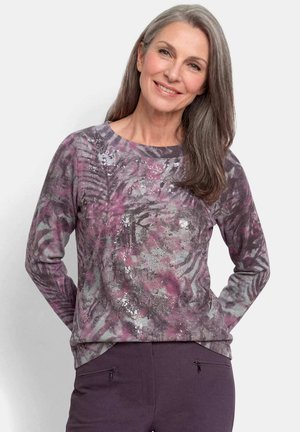 Frau mit langen grauen Haaren, die lächelt, trägt ein langärmeliges Shirt mit abstraktem Muster in Lila und Grau sowie dunkelviolette Hosen mit Reißverschlusstaschen.