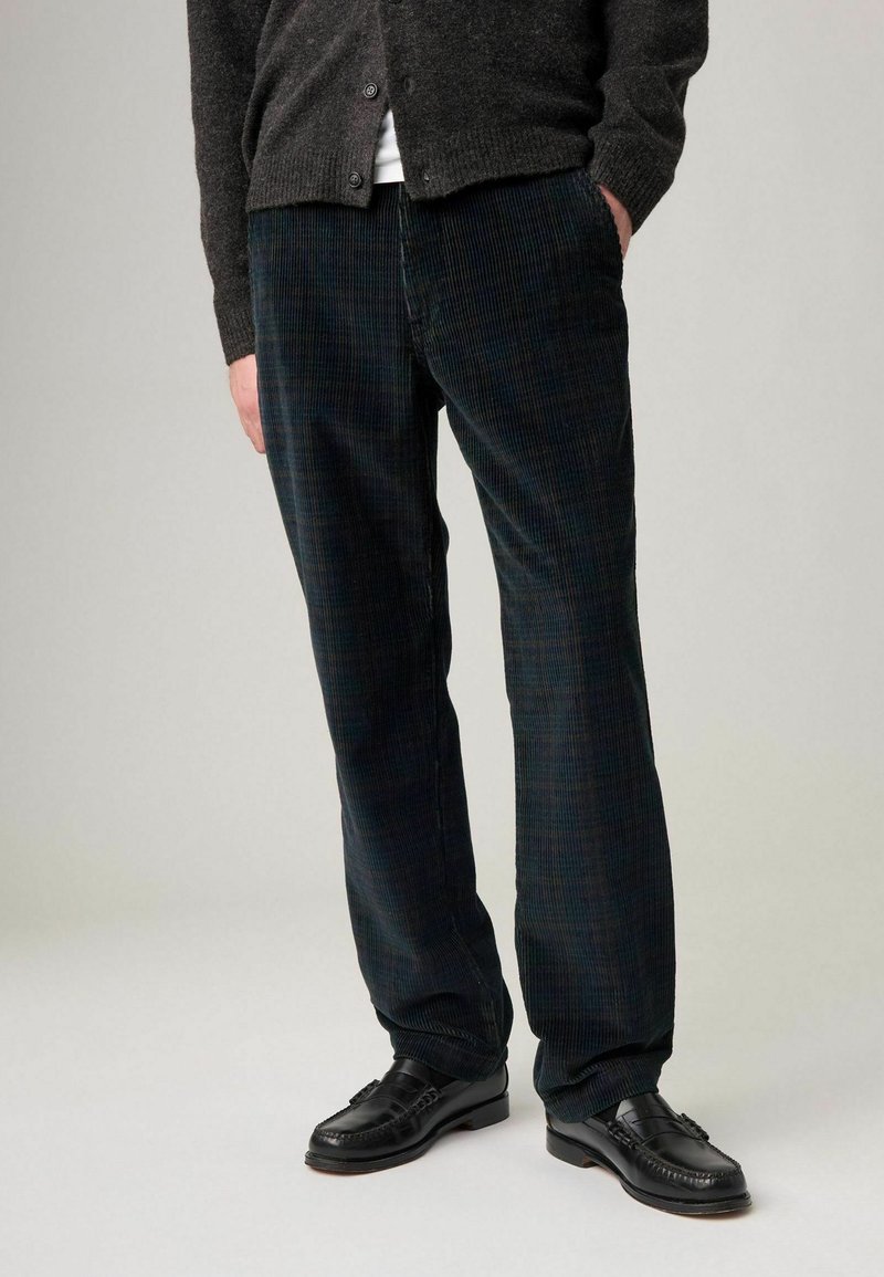 Dunkelgrüne Cordhosen für Herren mit einem dezenten Karomuster und geradem Bein. Kombiniert mit schwarzen Leder-Loafern.