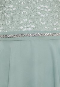Robe vert menthe avec corsage en dentelle, bordure en strass argentés à la taille et jupe en tissu lisse sous la bordure.
