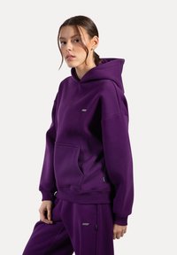 Sudadera con capucha morada, con un bolsillo frontal, puños y dobladillo acanalados. Presenta un pequeño logo en el pecho, confeccionada con un tejido suave.