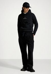 Μαύρο φούτερ με κουκούλα και κορδόνια, με μικρό λογότυπο, συνδυασμένο με μαύρο παντελόνι cargo. Το σύνολο ολοκληρώνεται με μαύρα αθλητικά παπούτσια και καπέλο.