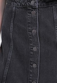 Zbliżenie na ciemnoszary dżinsowy spódnicę z widocznymi pionowymi szwami i centralnym rzędem metalowych nap. 