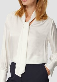 Blouse blanche à manches longues avec un détail de nœud au cou. Fabriquée en tissu doux et lisse, avec des poignets froncés. Portée avec un pantalon sombre.