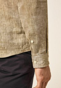 Chemise en lin de couleur marron clair avec une finition texturée. Les poignets boutonnés présentent des boutons blanc cassé et des détails de couture subtils.