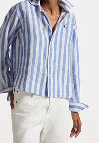 Linned skjorta med blå och vita vertikala ränder, button-down krage och liten logotyp på bröstet; matchad med vita byxor.