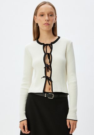 Cardigan blanc côtelé avec des finitions noires et des liens sur le devant, manches longues et coupe courte, associé à une ceinture noire.
