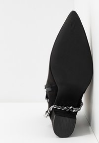Botte noire à bout pointu avec talon bloc présentant un détail de chaîne argentée autour de la cheville, vue de la semelle sur fond blanc.