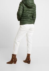 Zielona kurtka puchowa z kapturem, białe dżinsy oraz brązowe botki ze wzorem węża na kostkę z grubym obcasem. Teksturowe materiały i dopasowany krój.