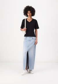 Maglietta nera in cotone a scollo a V, gonna maxi in denim blu chiaro con spacco sul davanti, sneakers bianche e una piccola borsa a spalla color crema.