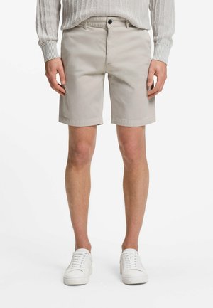 Mężczyzna ubrany w beżowe krótkie spodenki za kolano, jasnoszary dziergany sweter i białe trampki, stojący na tle jednolitego białego tła.