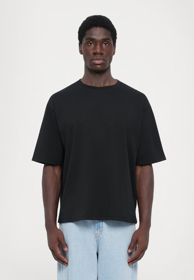T-shirt nera in cotone con spalle scese, vestibilità oversized, scollo rotondo e texture liscia; abbinata a jeans in denim chiaro.