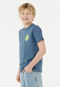 T-shirt en coton bleu avec un graphique rond néon vert sur la poitrine, manches courtes et encolure ras du cou. Porté avec un jean en denim bleu clair.