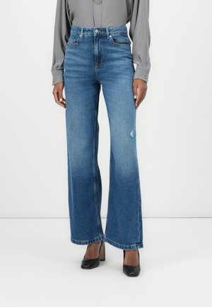 Personne portant un jean bleu taille haute à jambes larges associé à des chaussures à talons noirs et une chemise grise à manches longues.