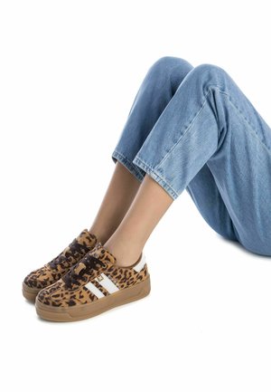 Zapatillas de leopardo con acentos en marrón y negro, que presentan tres rayas blancas, una suela de goma y cordones marrones. Pantalones vaqueros de color azul claro visibles.