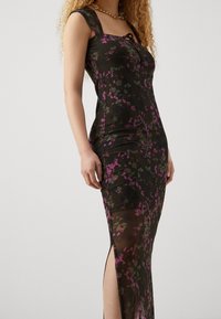 Blumenkleid in mittellanger Form aus transparentem schwarzem Stoff mit lila und grünen Mustern, figurbetonter Silhouette, quadratischem Ausschnitt und seitlichen Schlitzen.