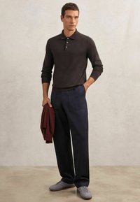 Dunkelbraunes langärmeliges Poloshirt mit drei Knöpfen, kombiniert mit marineblauen, weit geschnittenen Hosen. Graue Freizeitschuhe. Roter Pullover über einem Arm gelegt.