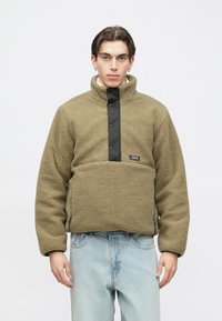 Fiatal férfi, olívazöld sherpa polárpulóvert visel fekete patentgombokkal és világoskék farmernadrágot, egyszerű fehér háttér előtt áll.