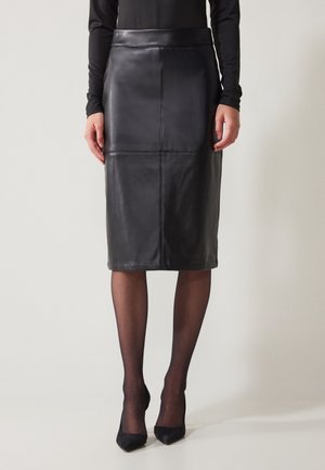 Femme portant une jupe crayon en cuir noire longueur genou, collants noirs transparents, talons aiguilles noirs et un haut noir à manches longues.