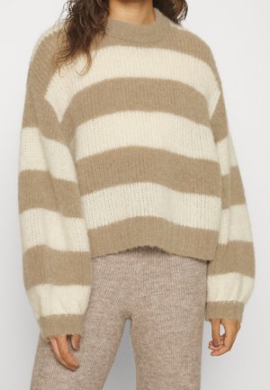 Personne portant un pull épais à rayures beiges et blanches, en tissu duveteux, à manches longues, et un pantalon en maille beige, debout devant un fond clair uni.