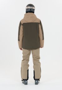 Giacca da neve in verde scuro e beige, abbinata a pantaloni beige. Presenta un cappuccio e zip, indossata con stivali da sci neri e grigi.