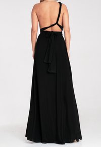 Femme portant une longue robe noire dos nu avec une bretelle torsadée sur l'épaule droite et une ceinture nouée à la taille.