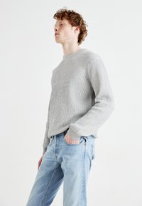 Pull en tricot côtelé gris avec col rond, associé à un jean en denim bleu clair. Texture marquée et coupe décontractée.