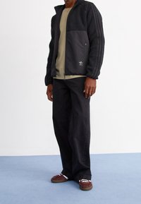 Black fleece and fabric zip jacket with textured sleeves, green t-shirt, black pants, and maroon shoes with laces.