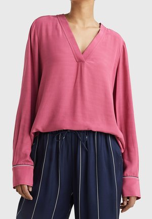 Femme portant un chemisier rose à manches longues et col en V, ample, rentré dans un pantalon bleu marine à rayures verticales blanches et taille à cordon.