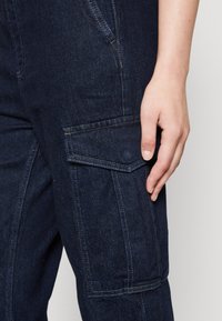 Marinblå denim cargo byxor med sidficka, som har kontrasterande sömmar och en avslappnad passform. Tyget har en något strukturerad yta.