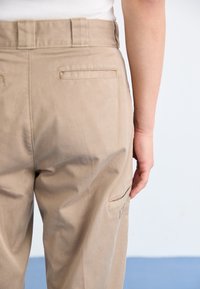 Pantaloni beige a vita alta, con tasche laterali e tasche posteriori. Tessuto liscio con leggere pieghe e dettagli cuciti sulle cuciture.