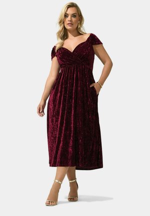 Mujer con un vestido midi de terciopelo burdeos con escote al hombro, mangas cortas y tacones altos dorados con tira en el tobillo, posando sobre un fondo liso.