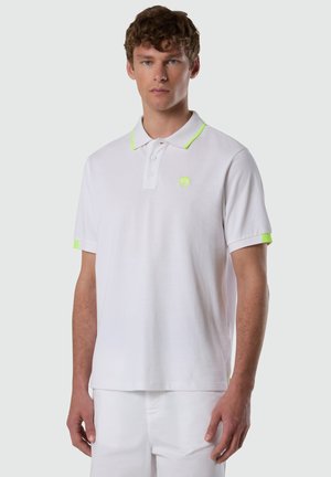 Giovane uomo che indossa una maglietta polo bianca con rifiniture neon verdi su colletto e maniche, in piedi davanti a uno sfondo chiaro e uniforme.