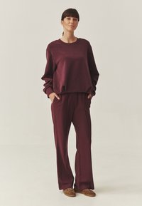 Bordeaux gebreide sweatshirt en bijpassende wijde broek, met lange mouwen, zijzakken en een relaxte pasvorm. Bruine suède schoenen maken de outfit compleet.