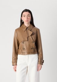 Een model poseert in een stijlvolle bruine leren jas met een kraag, gecombineerd met witte broek en oorbellen, tegen een effen achtergrond.