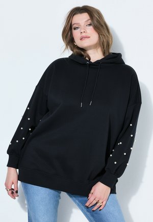 Młoda kobieta w czarnej bluzie z kapturem z perełkowymi ozdobami na rękawach i jasnoniebieskich dżinsach, stojąca na białym tle.