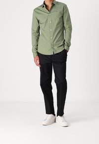 Camicia verde con bottoni e maniche lunghe, taschino sul petto, pantaloni neri e sneakers bianche su uno sfondo neutro.