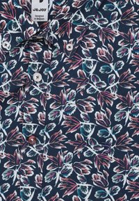 Marineblauwe shirt met bloemenpatroon in roze, groen en witte tinten, met een knoopsluiting en ronde kraag. Katoenen stof met een gladde textuur.