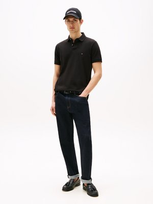 Polo noir, jean en denim foncé avec les poignets retroussés et mocassins noirs. Les accessoires comprennent une casquette noire avec un texte logo blanc.