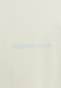 Lys beige bomulds T-shirt med en blød tekstur og et lyseblåt trykt logo, der står "Originals Studio Edition."
