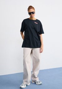Black cotton t-shirt with short sleeves and a small pink logo. Paired with loose beige trousers and multi-color athletic shoes.