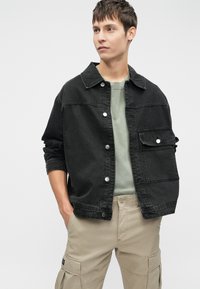Sort denimjakke med knaplukning, lomme på brystet og opsmøgede ærmer, båret over en grøn skjorte og parret med beige cargo-bukser.