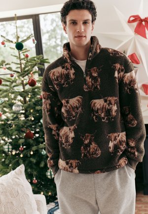 Un hombre luce un jersey polar marrón estampado junto a un árbol de Navidad decorado en un salón festivo.