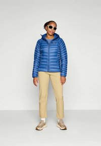 Vrouw in een blauwe pufferjack, beige broek, lichtbruine sneakers, witte sokken en witte zonnebril, staand tegen een effen lichte achtergrond.